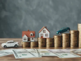 Miniatiūriniai automobilių ir namų modeliai yra šalia augančios pinigų krūvos.